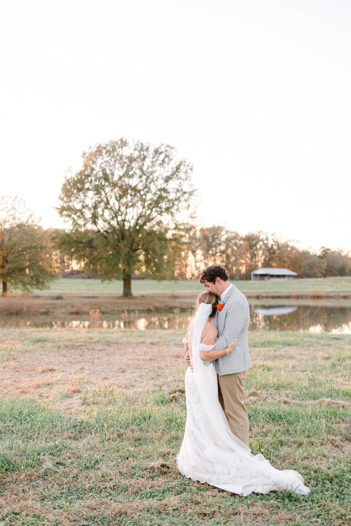 Bride and groom embrace in front of pond and tree at farm wedding near Charlottesville VA