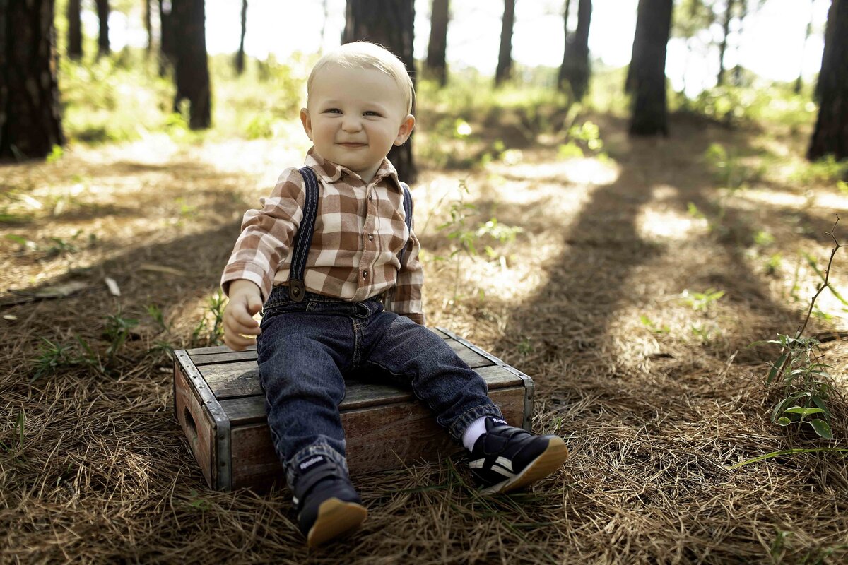 Cute birthday boy in suspenders and overalls, smiling brightly while surrounded by pine trees