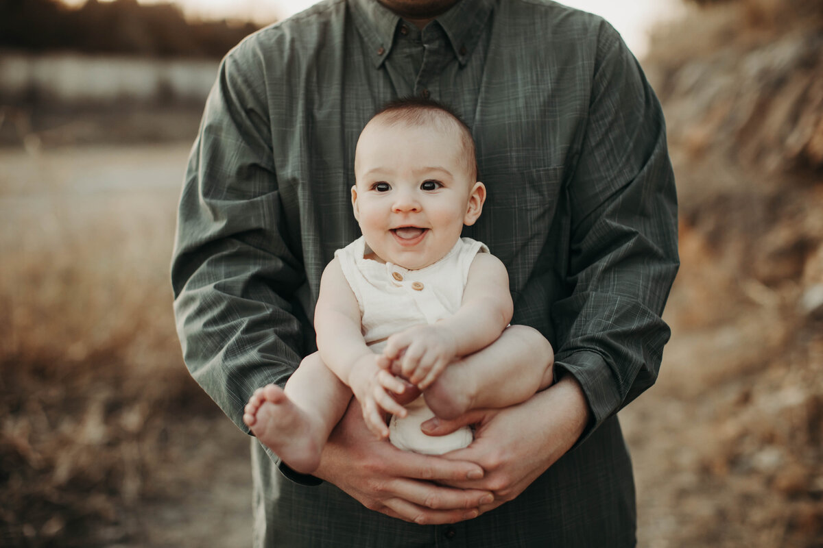Baby boy smiling big and holding his feet while dad holds him. 