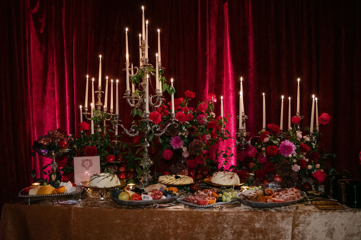 An elegant dining table setup with a luxurious candelabra filled with lit candles surrounded by vibrant red flowers. The table is adorned with various plates of appetizers and fruits, set against a rich red curtain backdrop, creating a warm and intimate ambiance.