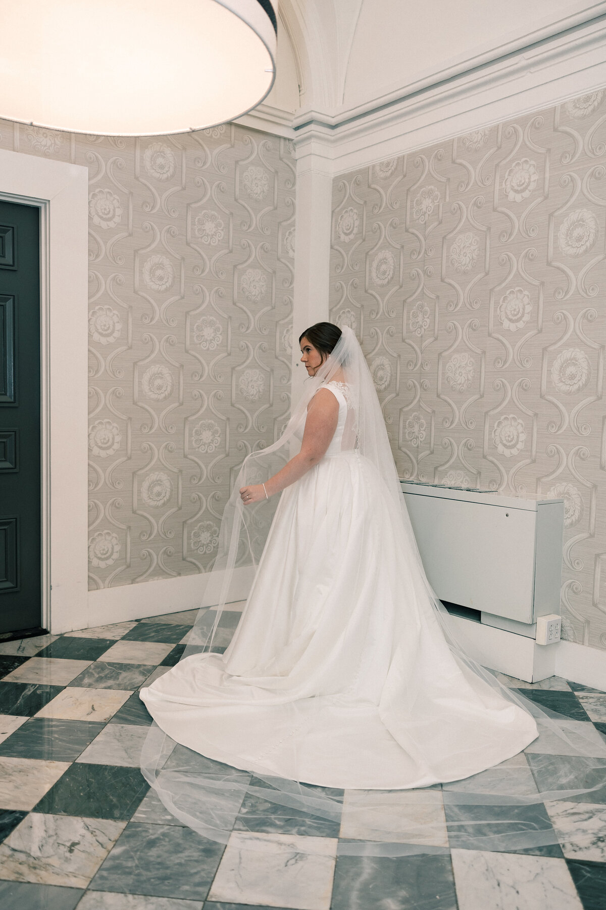 Bride in satin ballgown with cathedral veil twirling in Hotel Monaco bridal suite with marble floors and wallpaper.