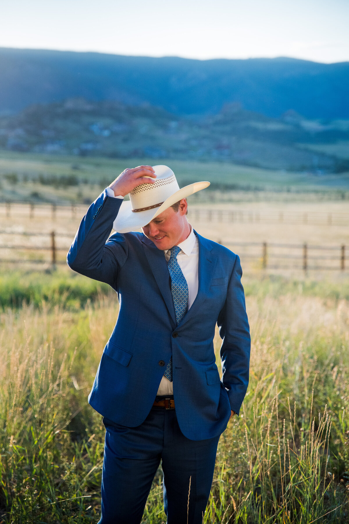 A groom tips his cowboy hat toward the camera standing in a field at his Colorado home.
