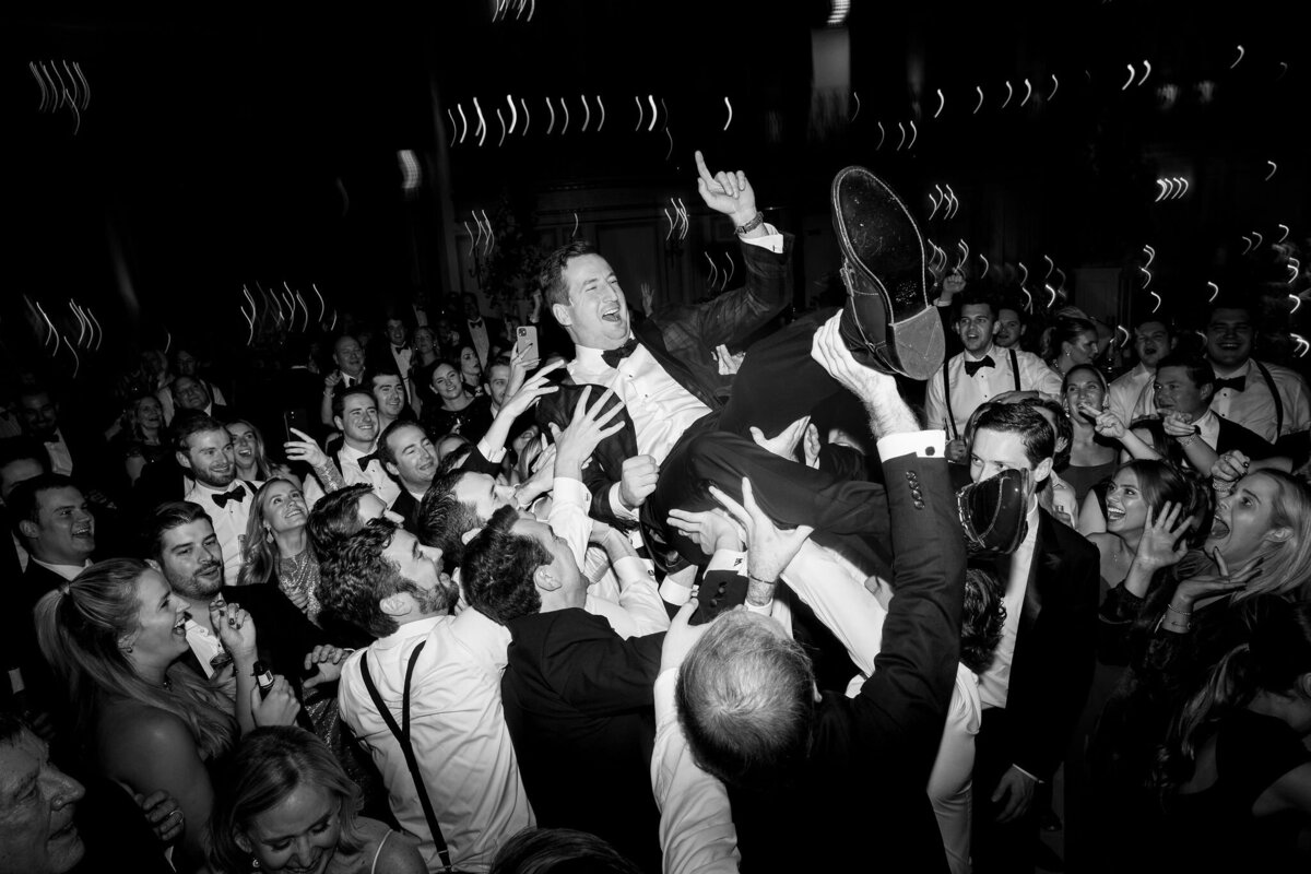 A picture of the groom being held up by the wedding guests. 