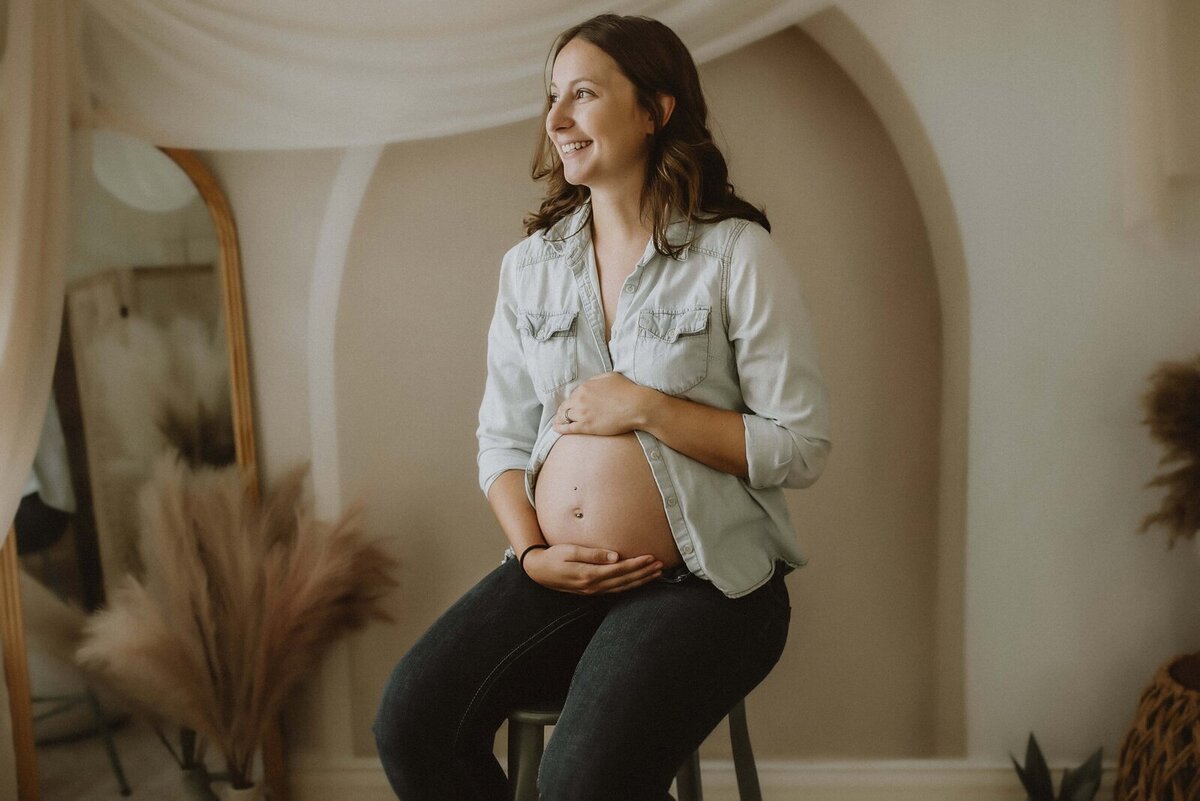 Pregnant woman smiling, seated with hands on belly, wearing a light denim shirt and dark pants, in a softly lit room with a mirror and decorative plants.