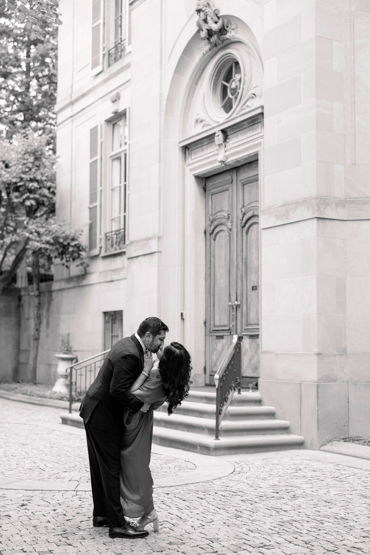 A couple shares a romantic kiss on a cobblestone path in front of an ornate building with tall windows and a grand wooden door. They are surrounded by trees, and the scene has a classic, timeless feel in black and white.