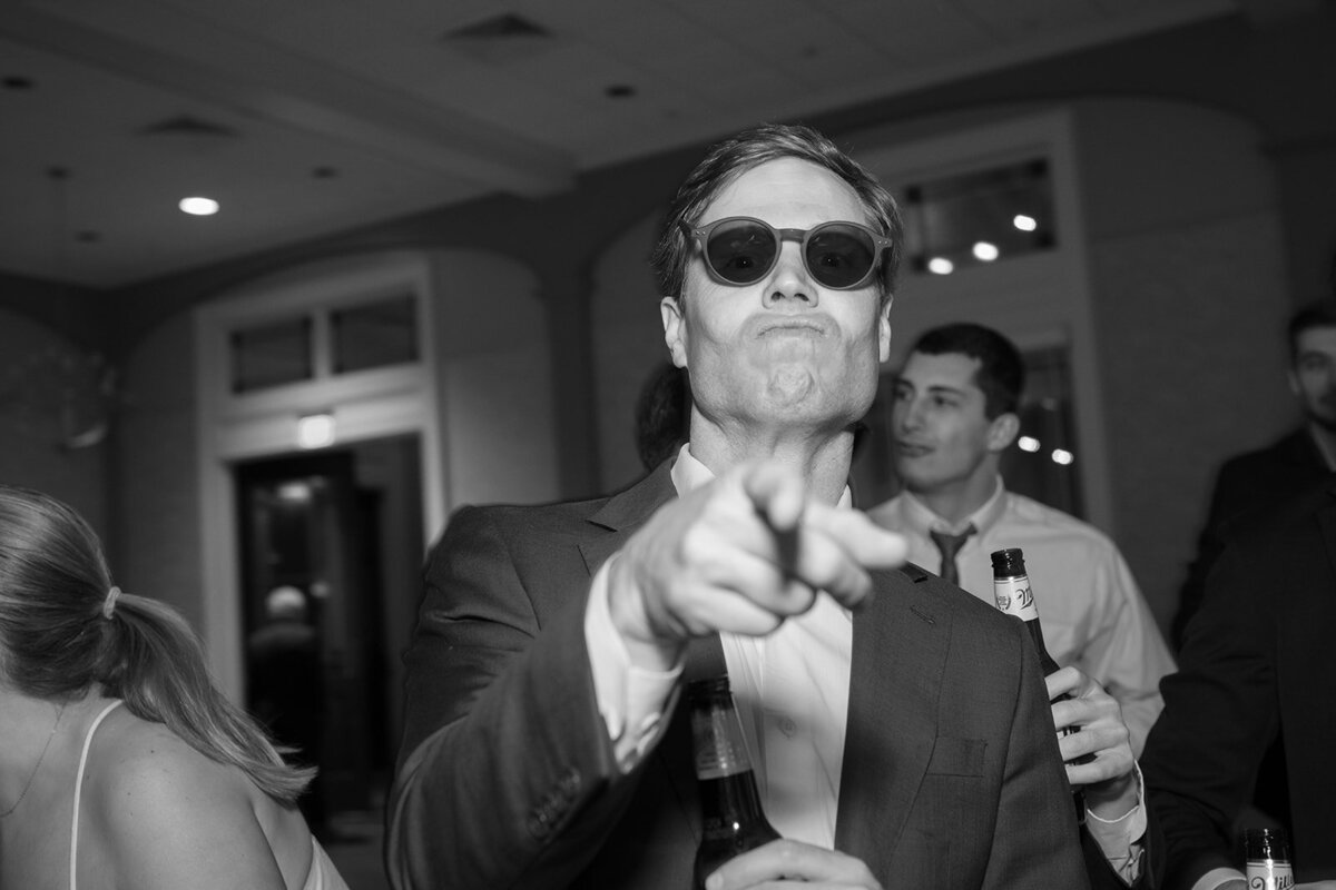 Black and white photo of a wedding guest in a suit and sunglasses pointing directly at the camera while holding a beer, taken during Callie & Hayes’ reception at Burritt on the Mountain.