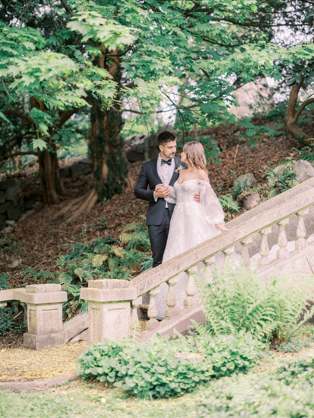 A couple in formal wedding attire stands on a stone staircase in a lush, green garden, expertly arranged by Destination Wedding Planner Melissa Dawn Event Designs. The groom wears a dark suit and the bride is in a white gown with a long train. They are holding hands.