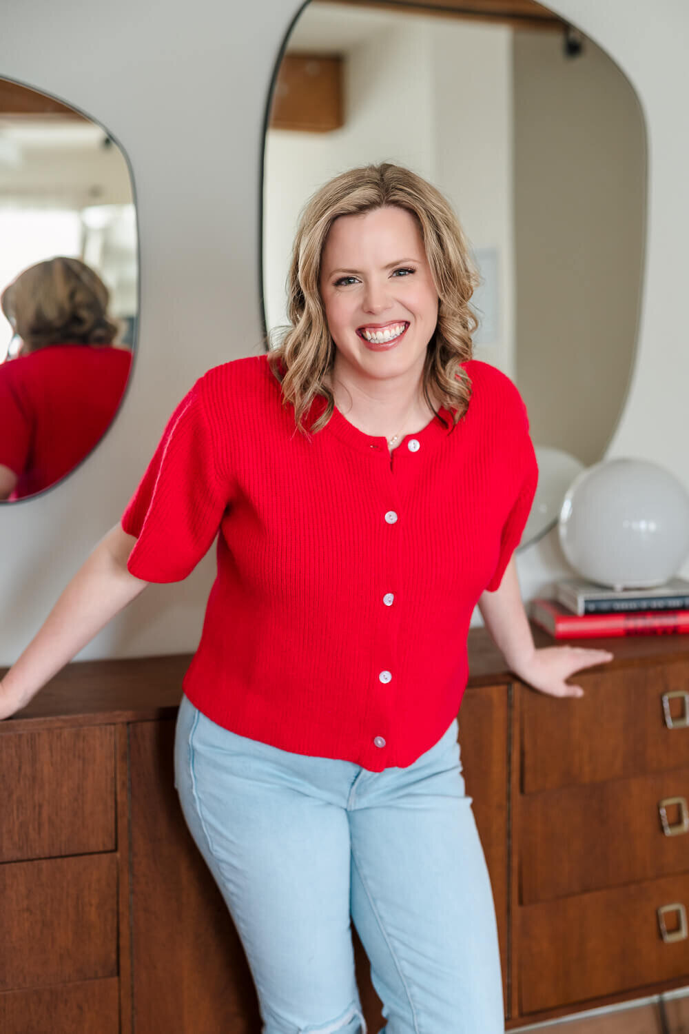  Women’s health specialist leaning against wooden console, smiling in red knit top and light jeans.