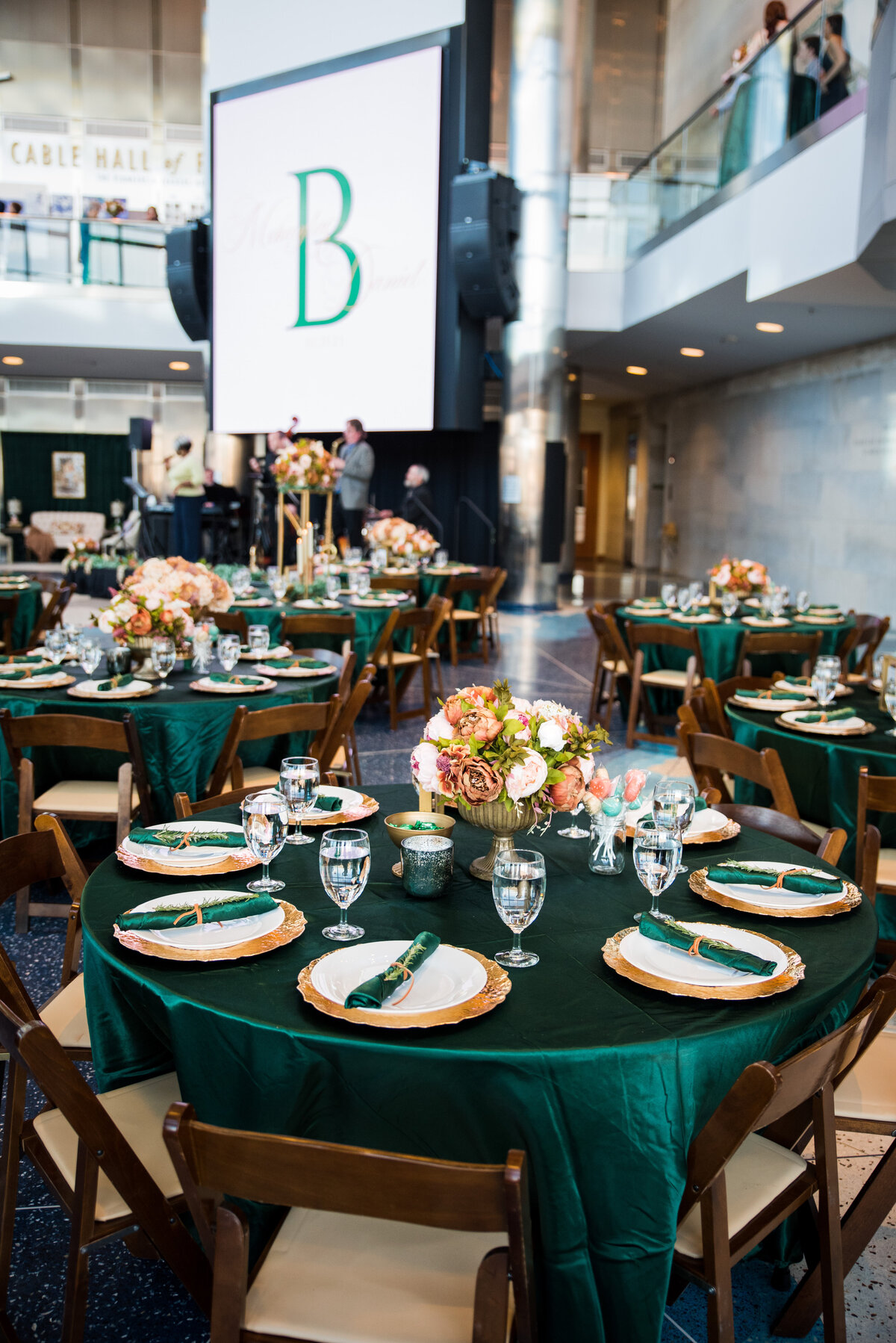 A round reception table is adorned with emerald green tablecloth, plates, and napkins. A large screen with the monogram letter "B" is in the background.