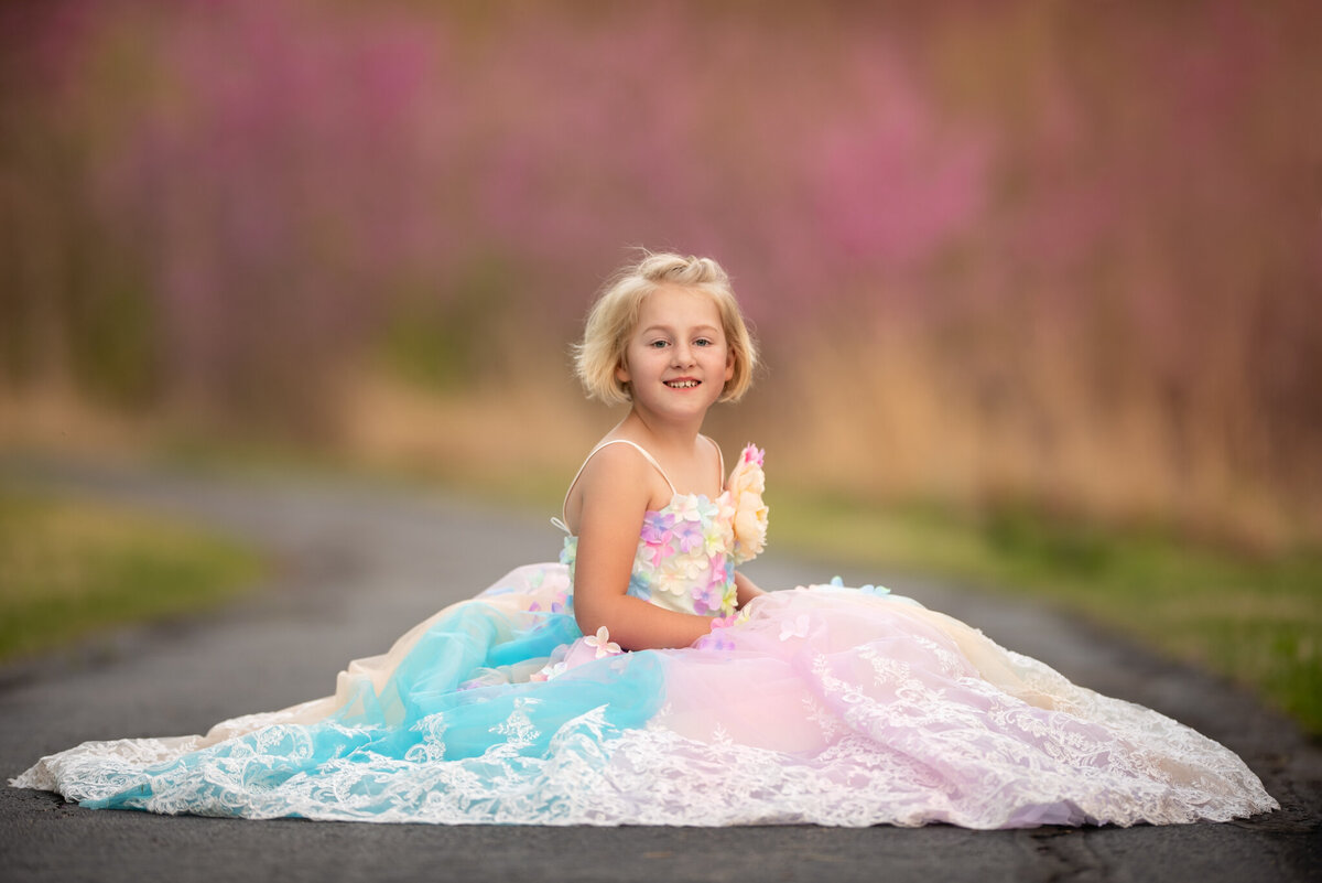 Sweet portrait of a young girl in a pastel pink and blue gown sitting in a sunlit meadow. Captured in Olathe, Kansas, this dreamy setup is ideal for families seeking colorful and outdoor child photography