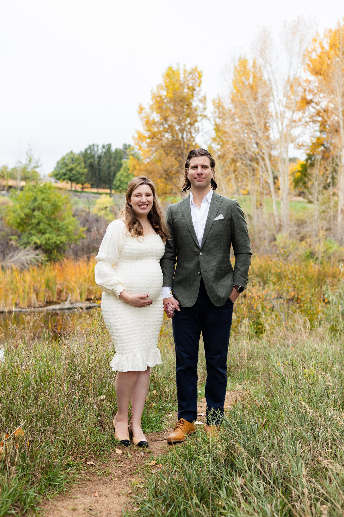 Pregnant woman and her husband hold hands and smile at the camera.