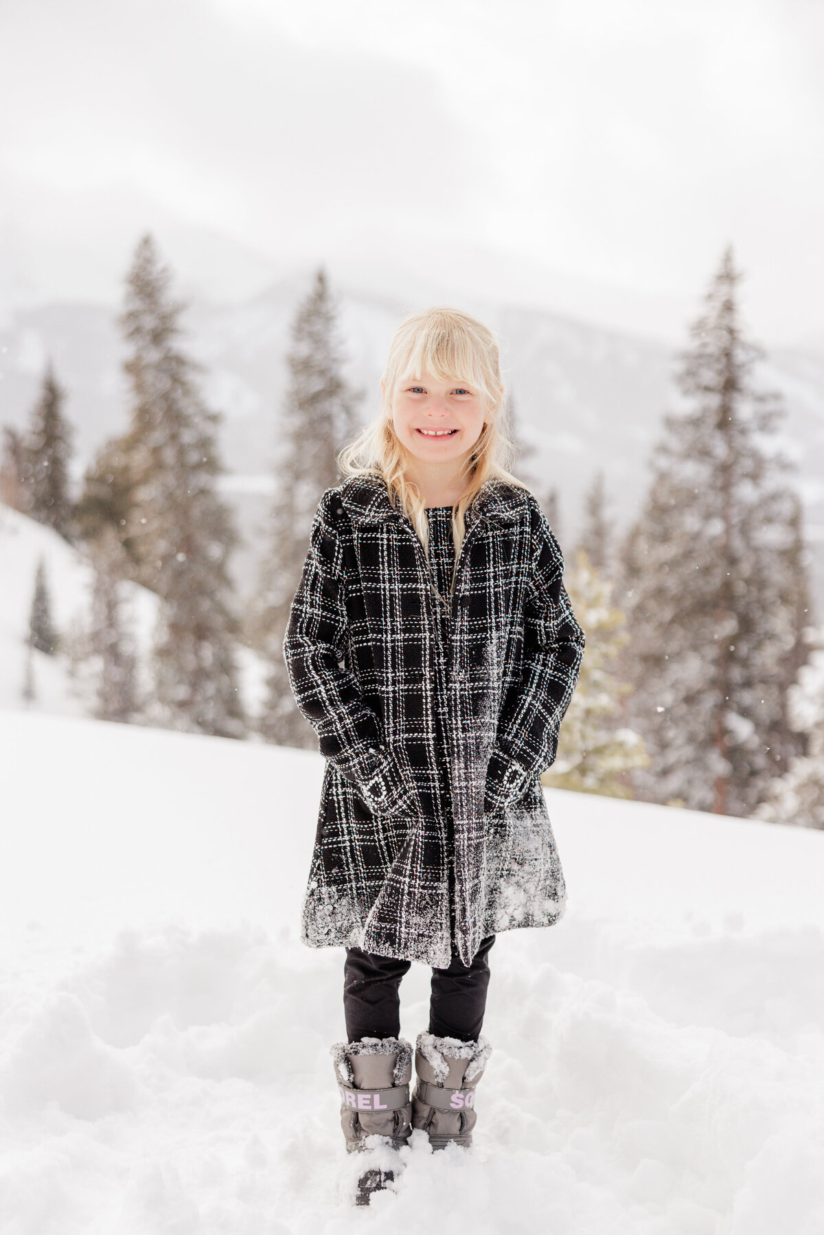 A young girl in a plaid coat smiles while standing in deep snow for her Breckenridge Family Photographer