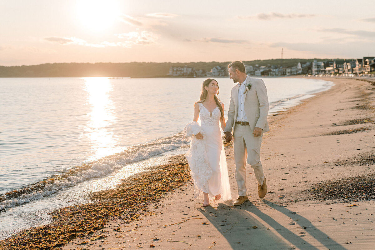 Newlywed couple walking hand in hand along the Connecticut shoreline, with the gentle waves and beach houses in the distance