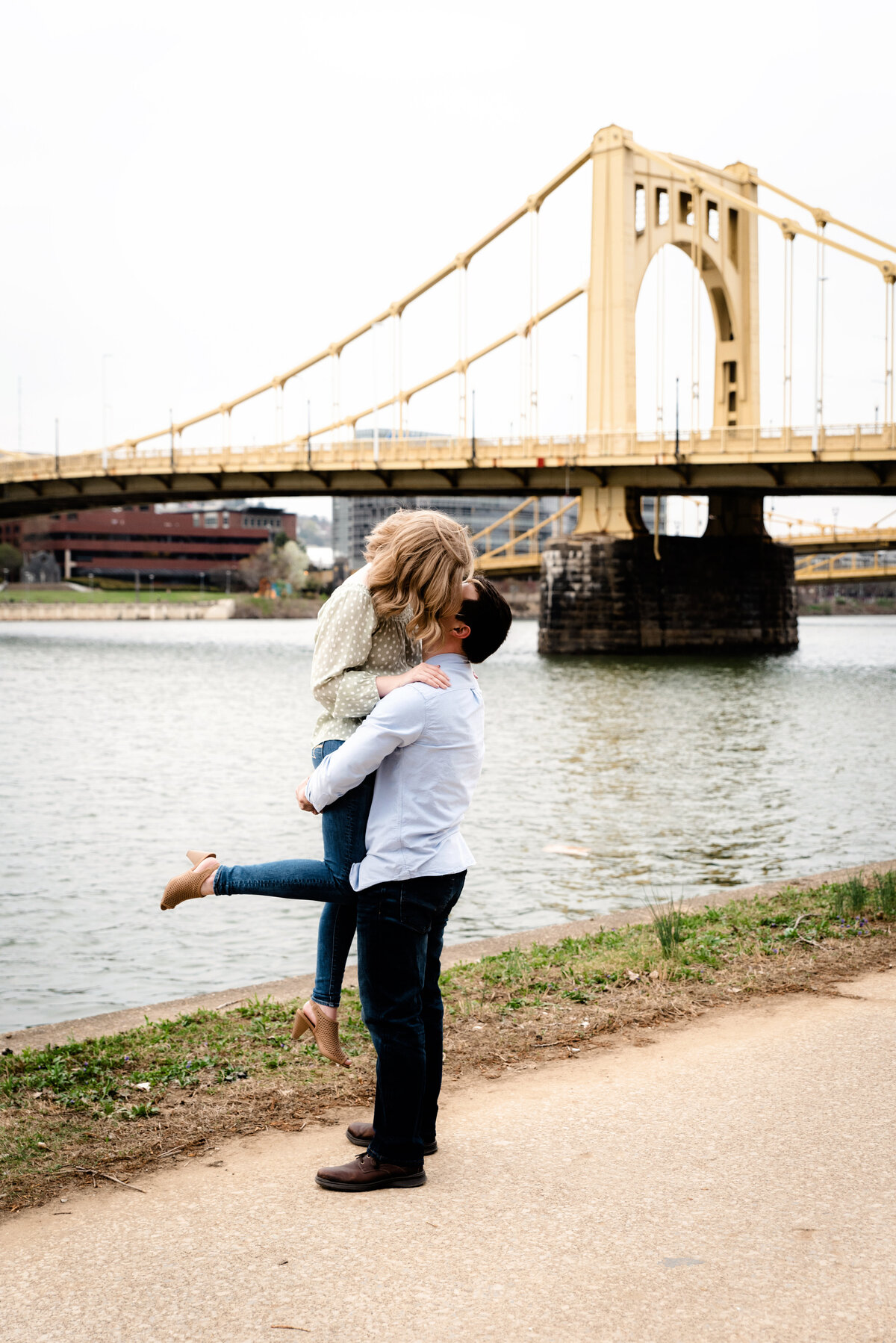 groom lifts bride up for kiss by river with roberto clemente bridge in background