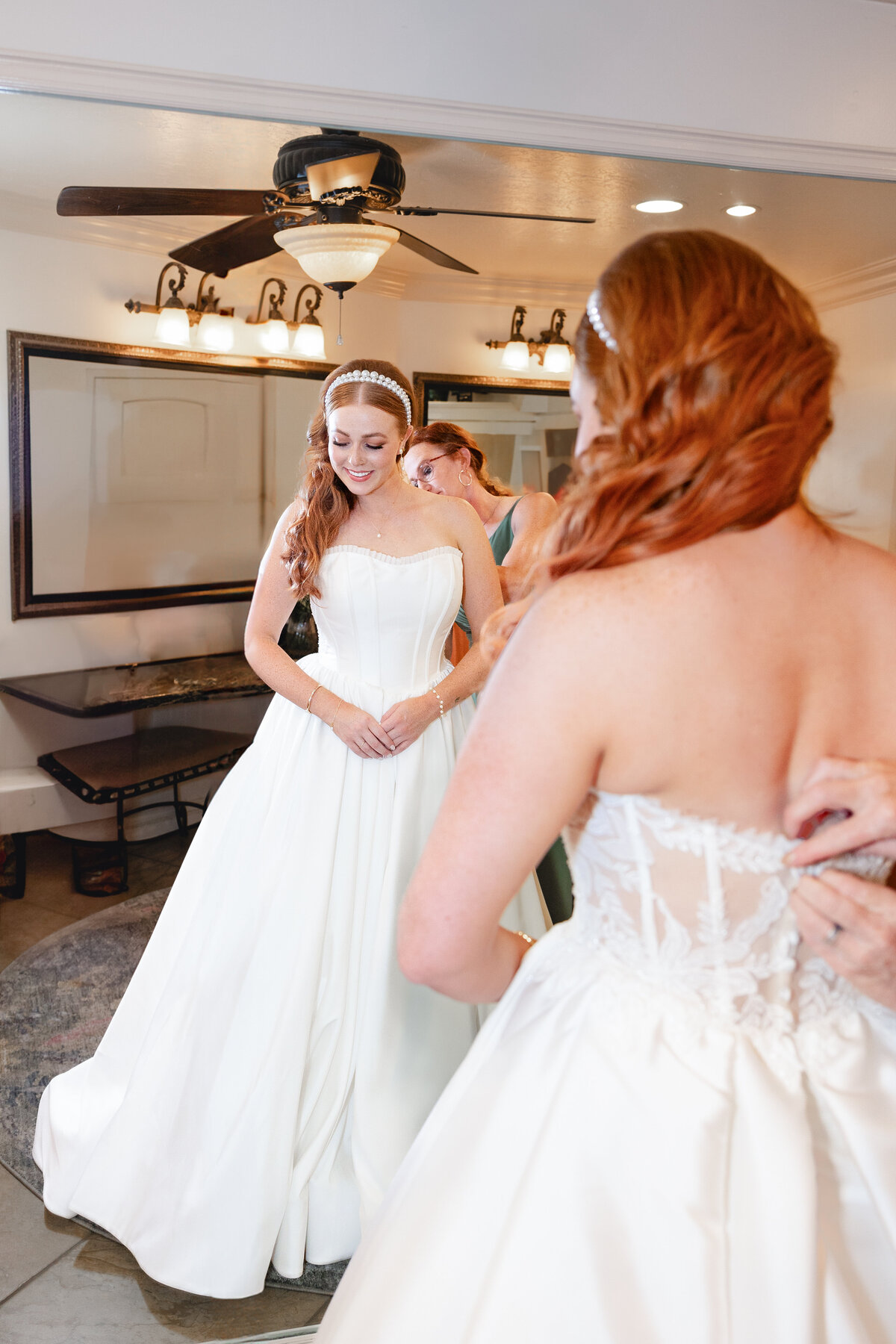A bride with red hair and a pearl headband smiles as she looks in the mirror, while someone adjusts the back of her wedding gown.