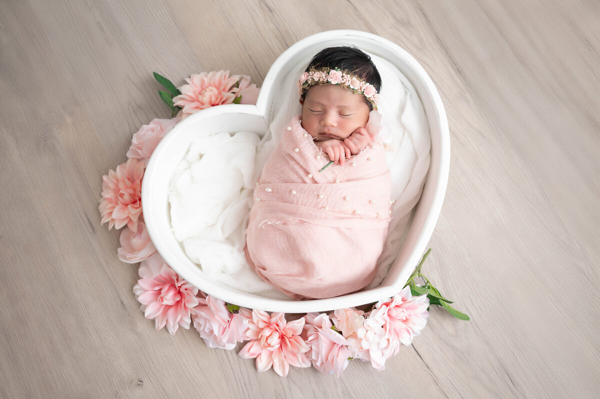 Newborn baby swaddled in a light pink wrap with delicate pearl accents, nestled inside a white heart-shaped prop surrounded by an arrangement of pink flowers.