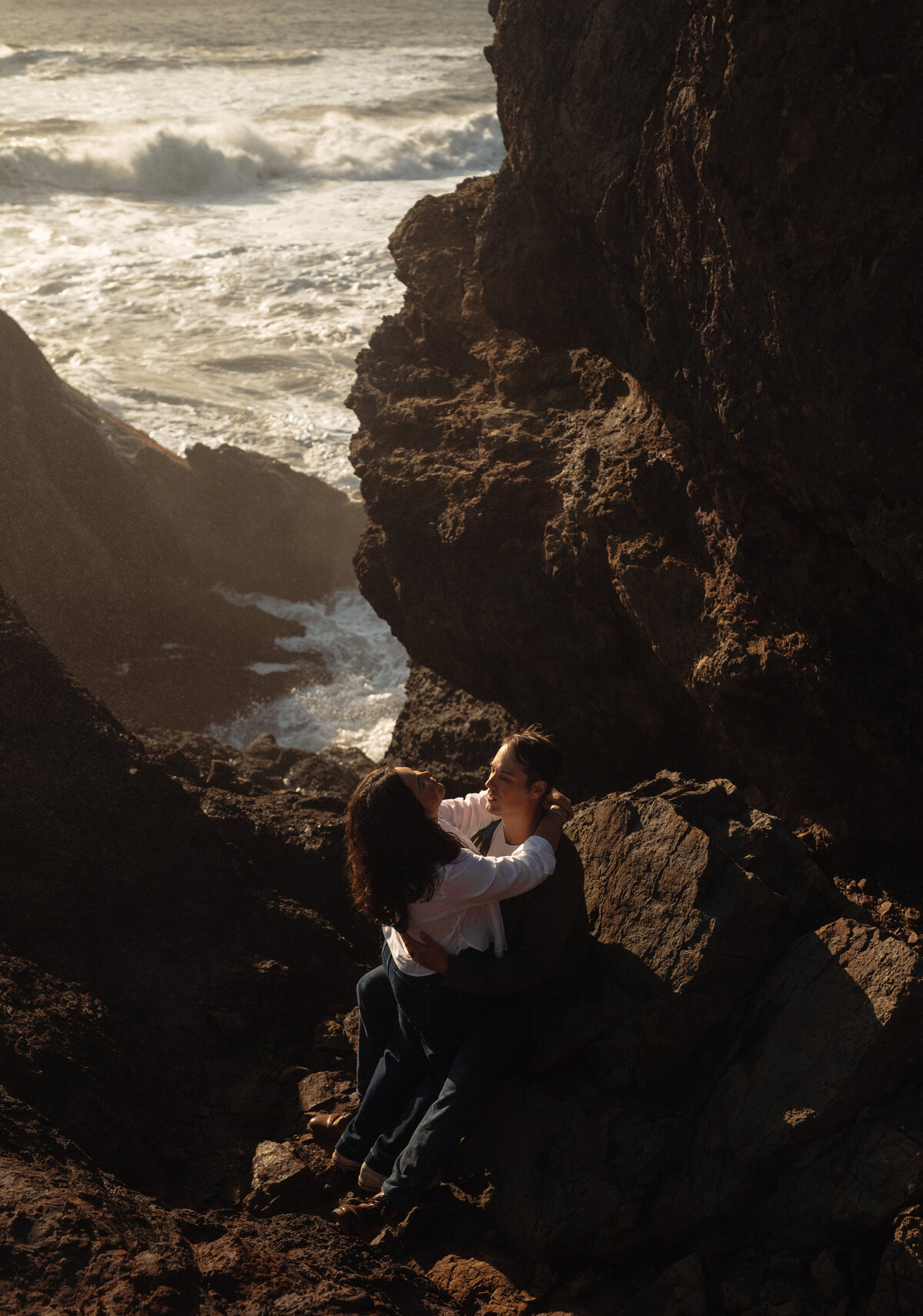 romantic moment of engaged couple smiling and holding each other with sunlit waves in the background