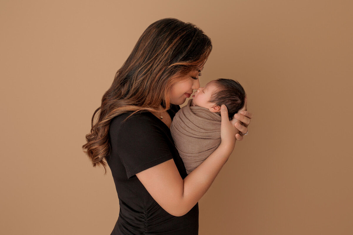 A mother gently holding her swaddled newborn baby close to her face, both wearing black clothing against a warm beige background.