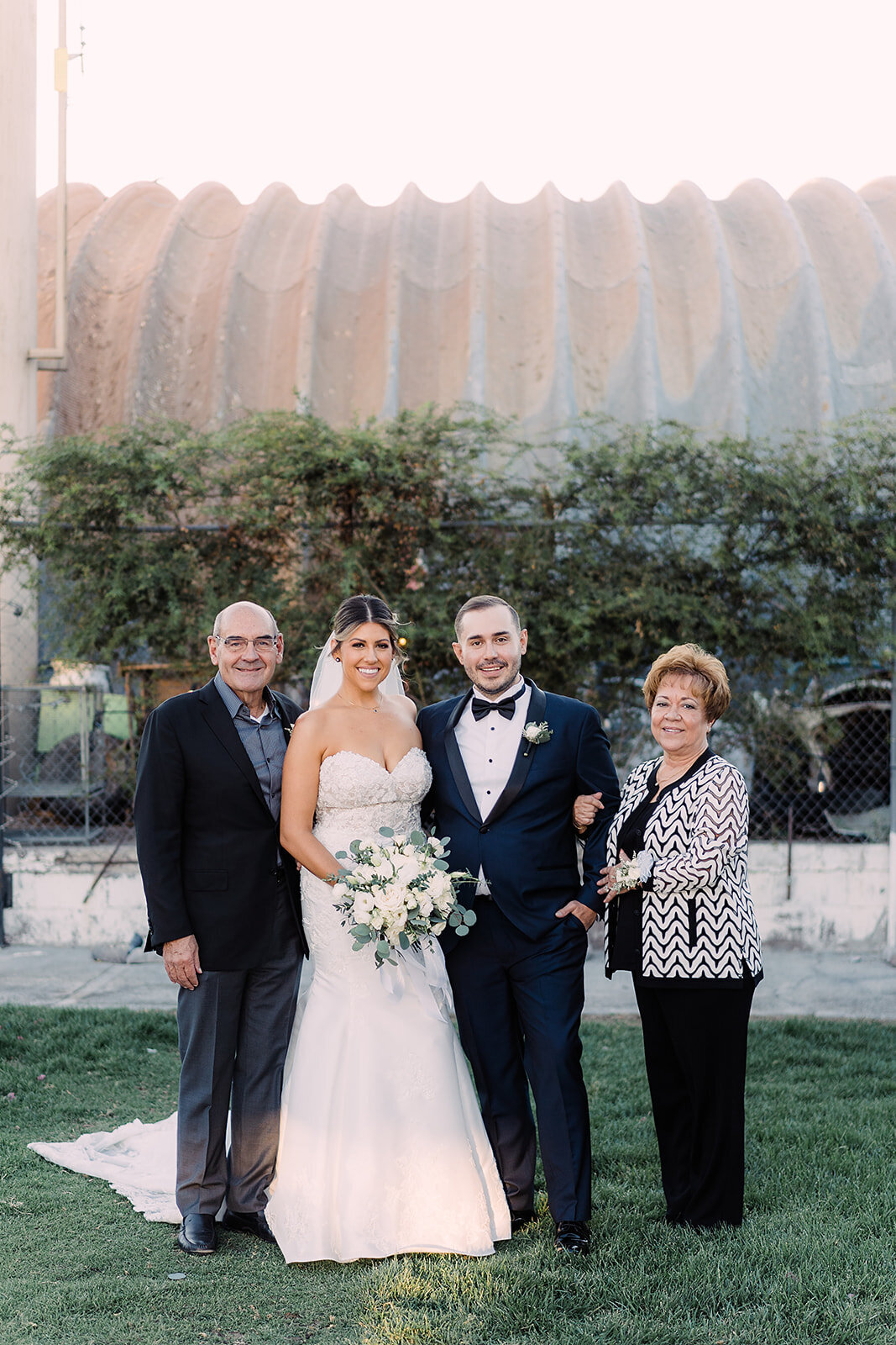 The couple stands outside with the groom’s parents, posing together on a green lawn with smiles, the bride holding a white floral bouquet.