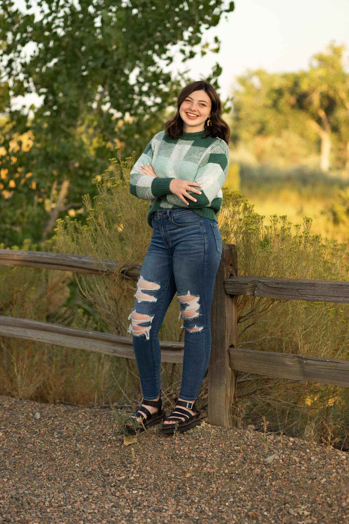 teenage girl wearing a green sweater and jeans leaning against a wooden fence on a trail