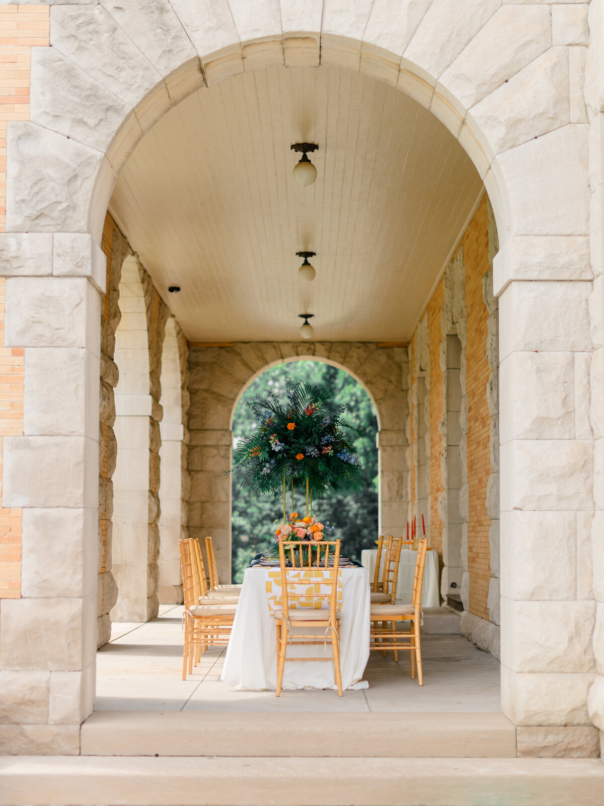 Elegant outdoor reception setup beneath the stone archways of Cairnwood Estate, featuring a bold floral centerpiece with lush greenery and vibrant orange blooms. Captured by Elizabeth Kane Photography, a Philadelphia Fine Art Wedding Photographer, this timeless al fresco tablescape blends historic architecture with sophisticated East Coast wedding inspiration.