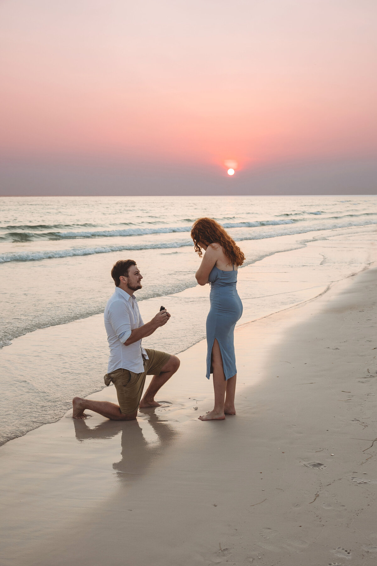Man kneels down on  the beach at sunset holding a ring box. Redheaded girl in blue dress covers her mouth in surprise.