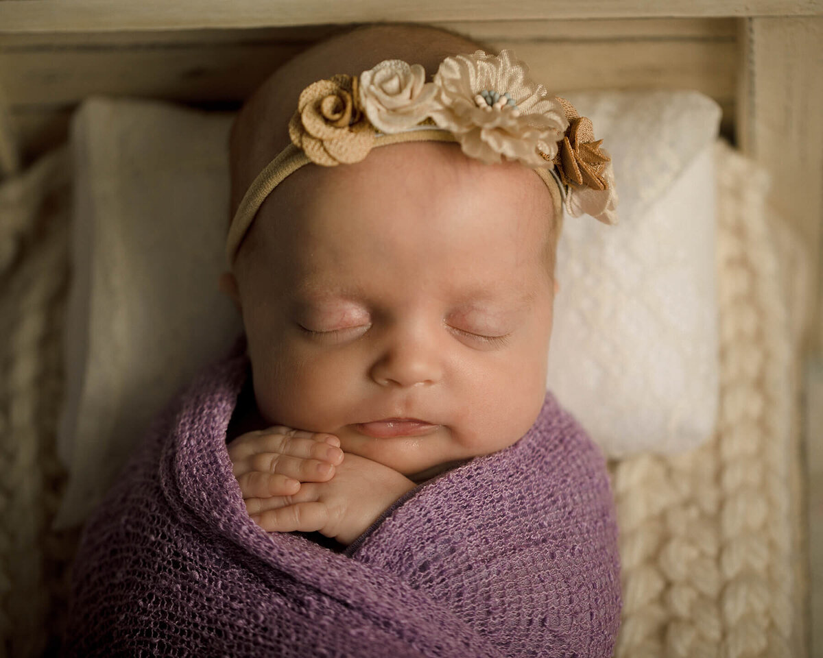 Newborn girl with purple wrap and flower headband laying in a small creme colored wooden bed.