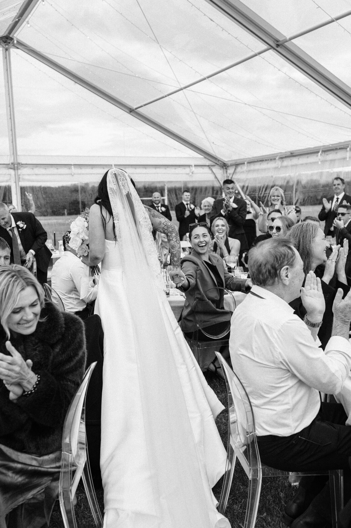 candid black and white photo of wedding guests cheering while bride walks through clear marquee in christchurch
