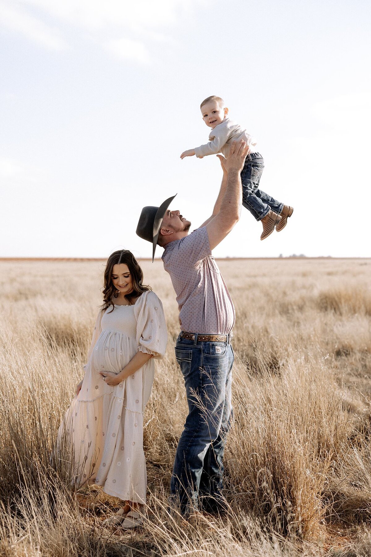maternity photo in open grass field near Lubbock Texas, dad tossing son in air and catching him, pregnant mom holding her belly