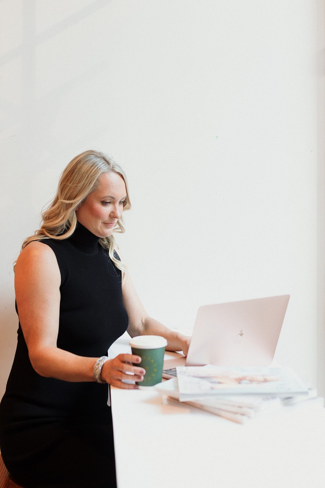 blonde woman sitting at a desk working on her laptop and drinking coffee. White background