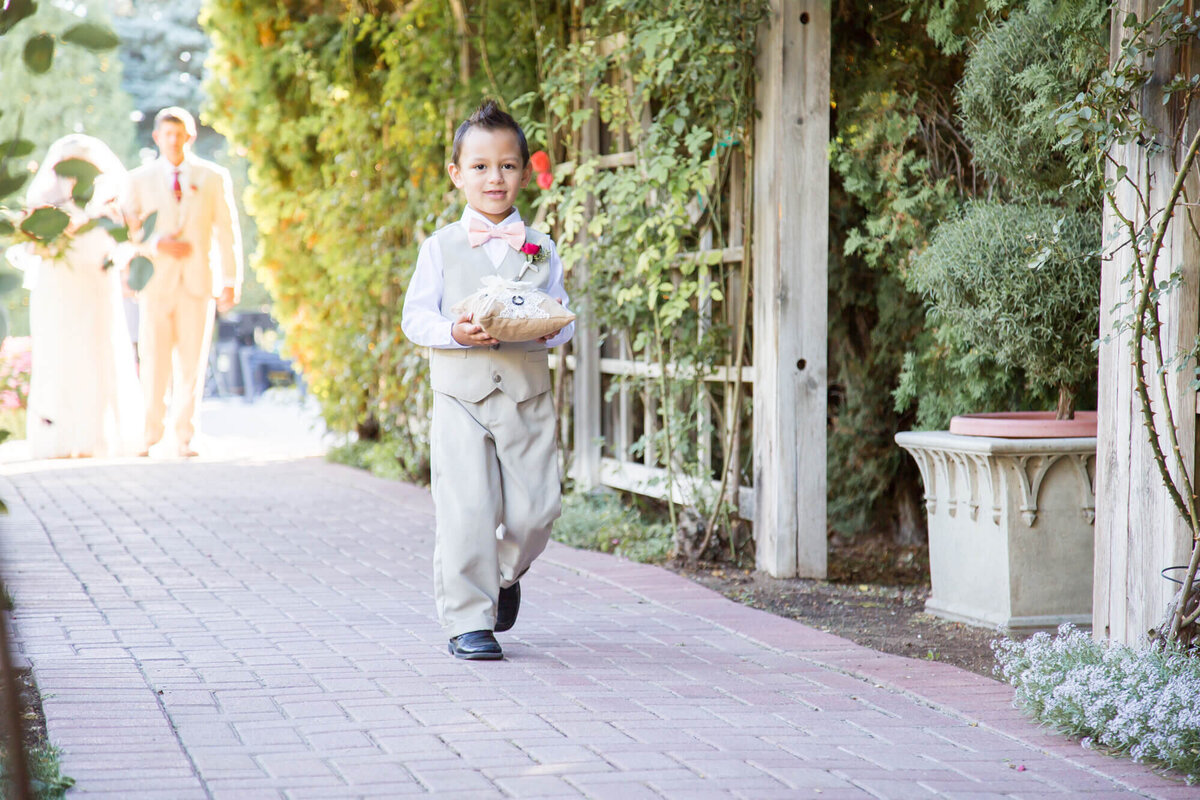 ring bearer in a cream vest carrying wedding rings to a classic garden wedding ceremony