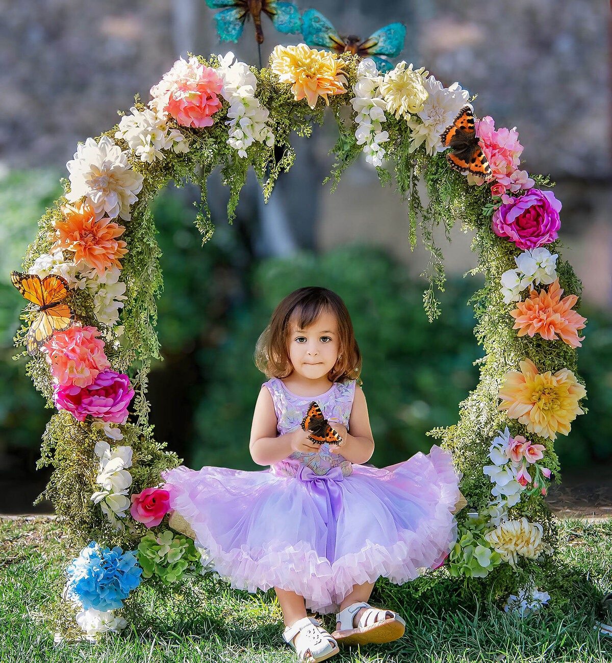 A young girl in a purple dress sits in a flower ring holding a butterfly for an Orange County milestone photography