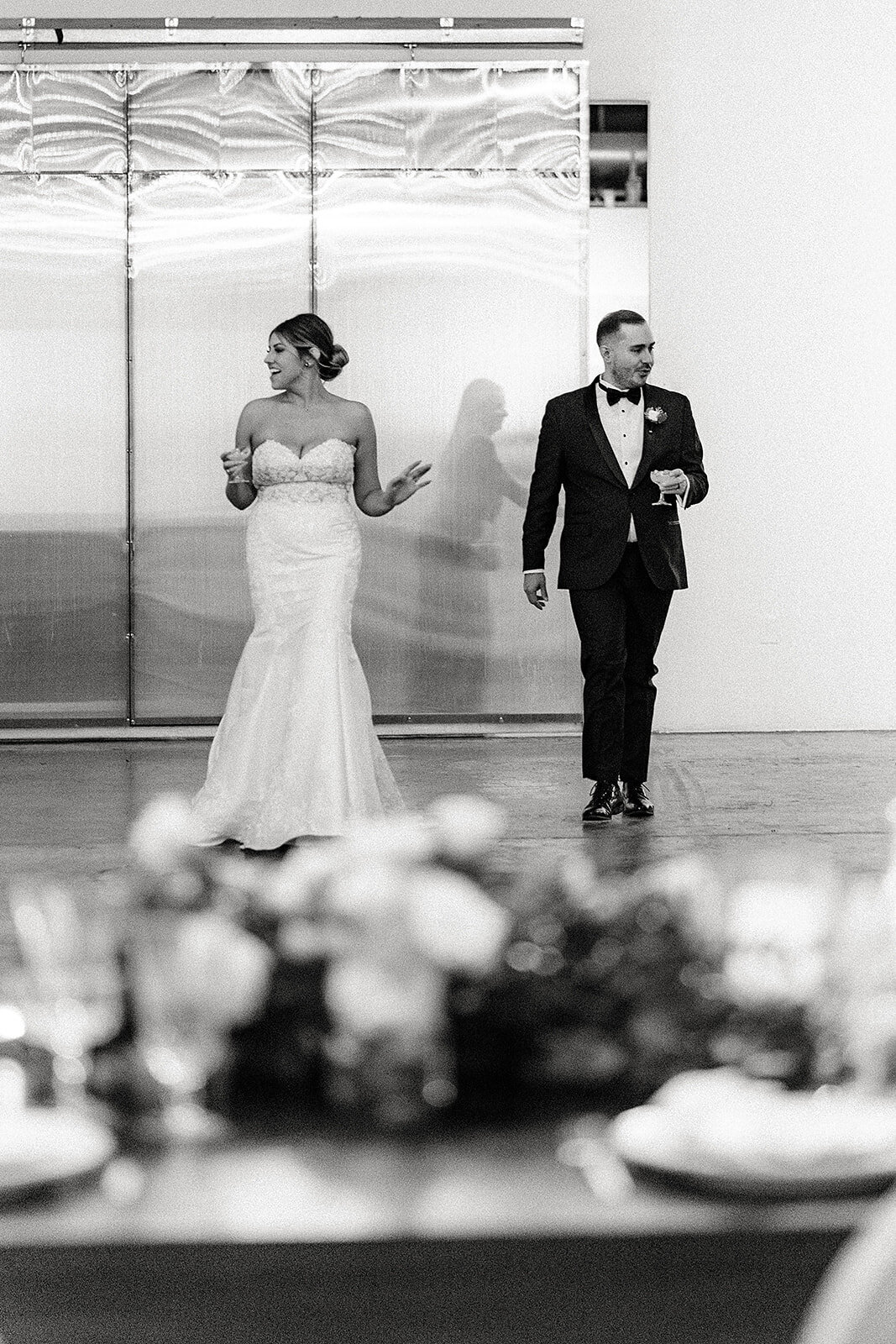 In a black-and-white shot, the bride and groom are seen dancing, with the bride holding a drink and smiling. They both look relaxed, enjoying a moment together on the dance floor.