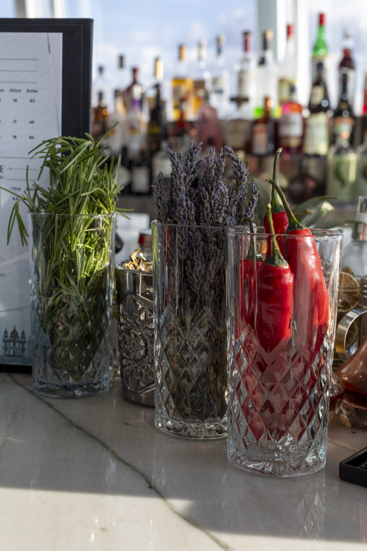 Garnishes on a bar sit in jars waiting for their moment to shine. Pictured are chili peppers, lavender, rosemary, and dried fruit in front of a fully stocked bar.