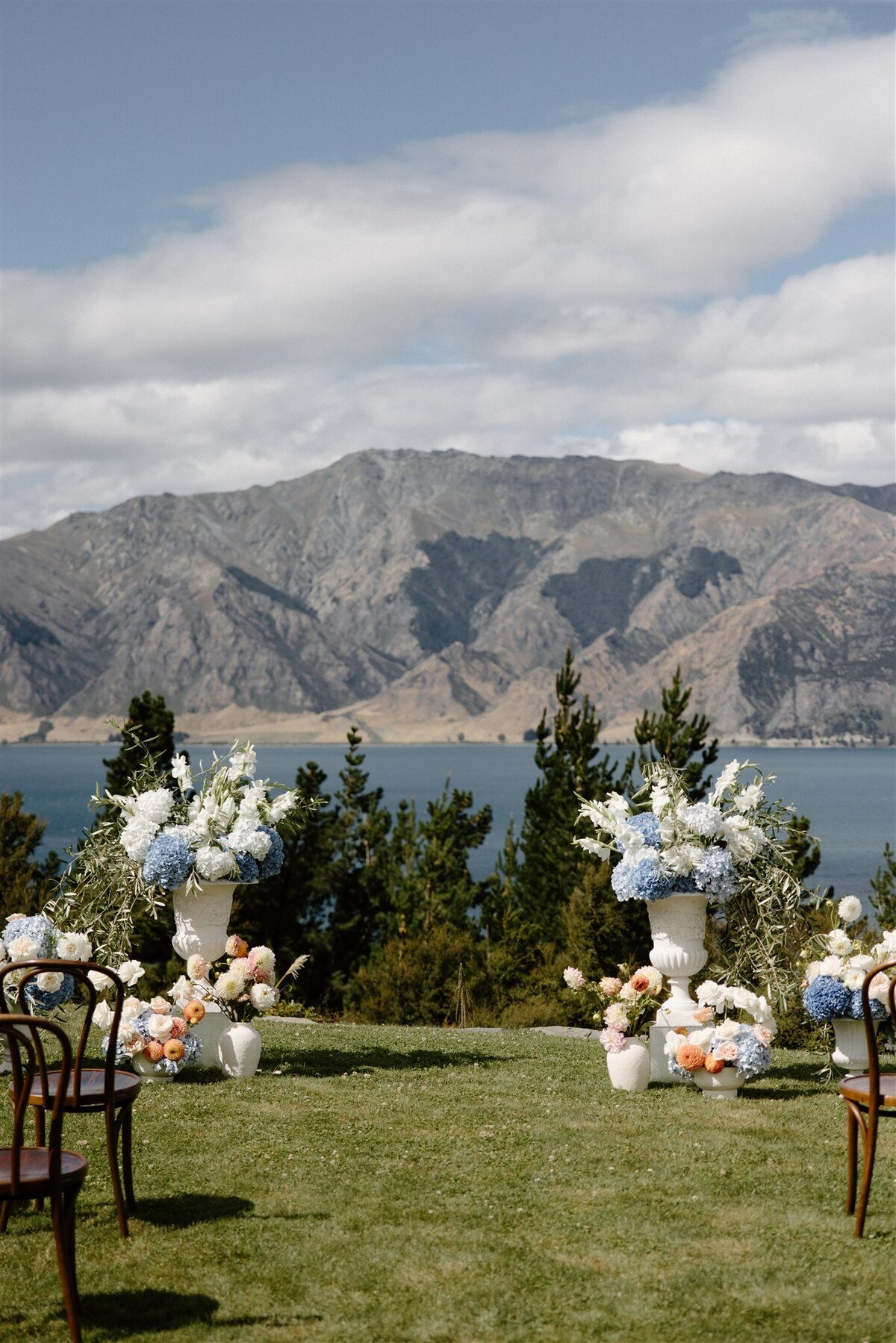 Stunning floral ceremony setup overlooking Lake Hawea, with large white urns filled with hydrangeas and roses, and mountain views in the background.