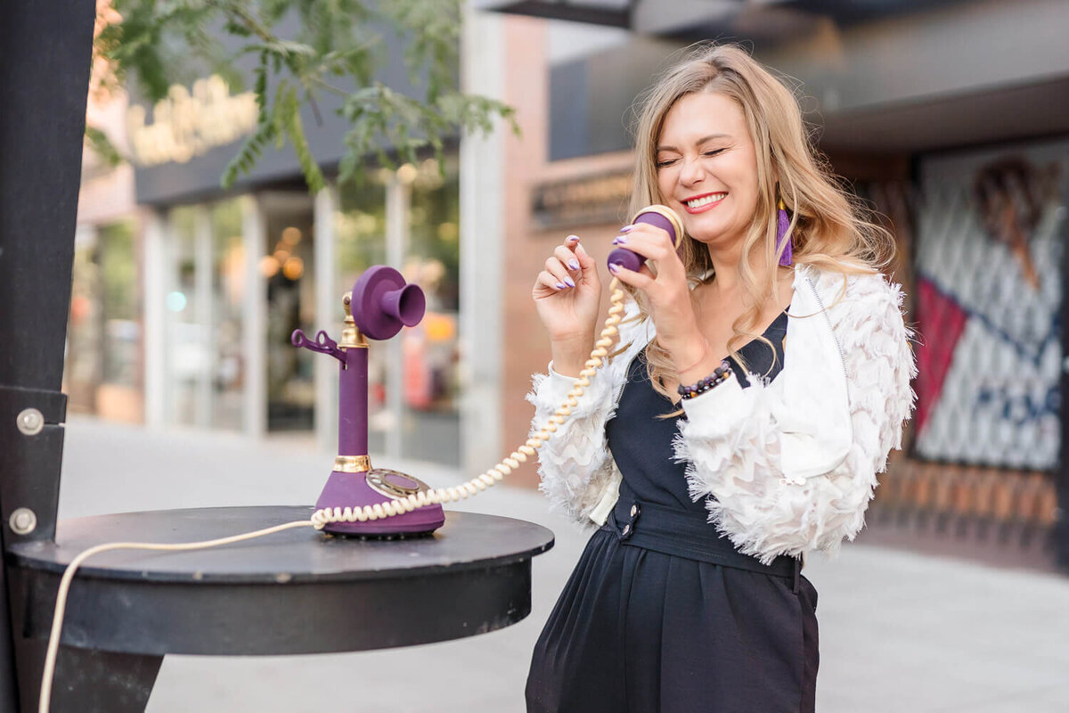 Female entrepreneur smiling while talking into vintage purple rotary phone in urban setting.