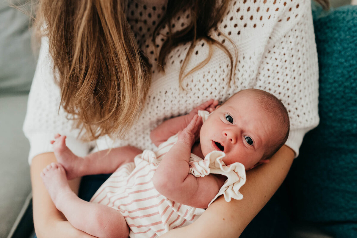 A mother wearing a white sweater holds her newborn daughter on the couch, the baby is looking directly at the camera and wearing a cute white and pink striped romper