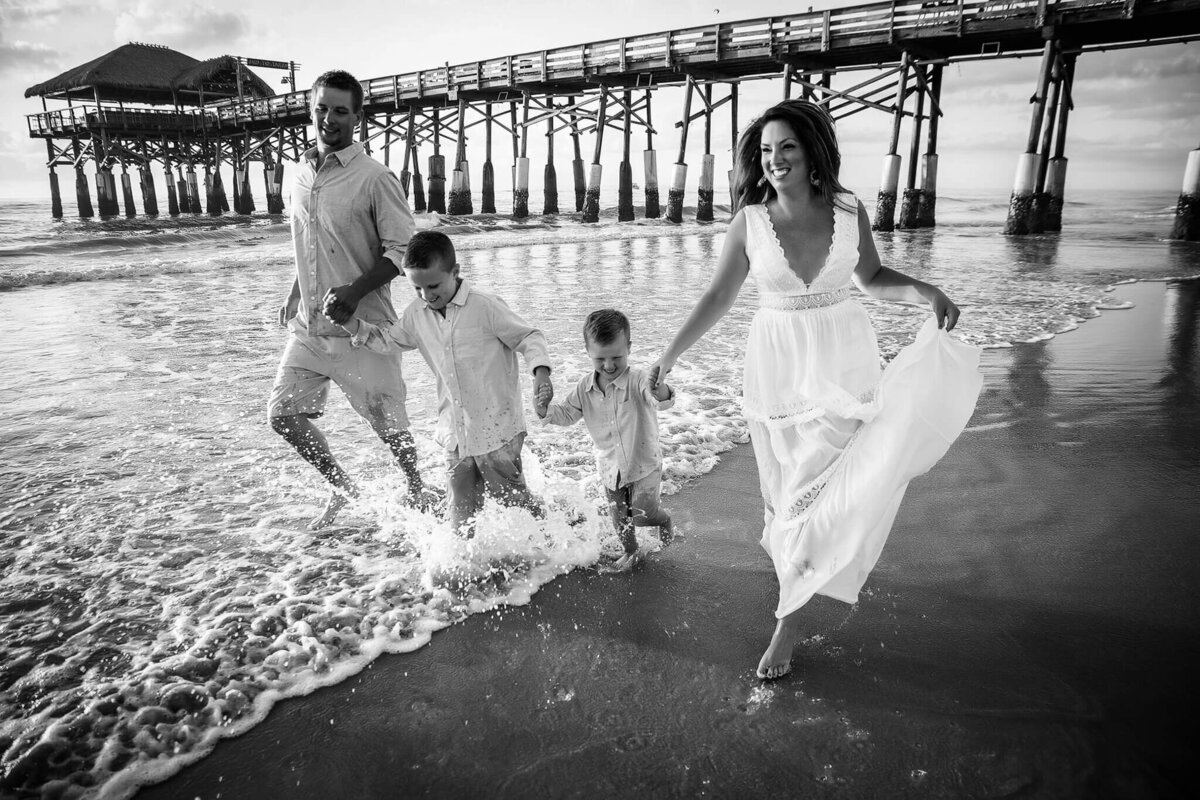 A family walks along Cocoa Beach, hand in hand, with waves splashing around their feet. The parents and two young children are smiling and dressed in light clothing. A wooden pier extends into the water in the background. Black and white photo capturing a serene Cocoa Beach session.