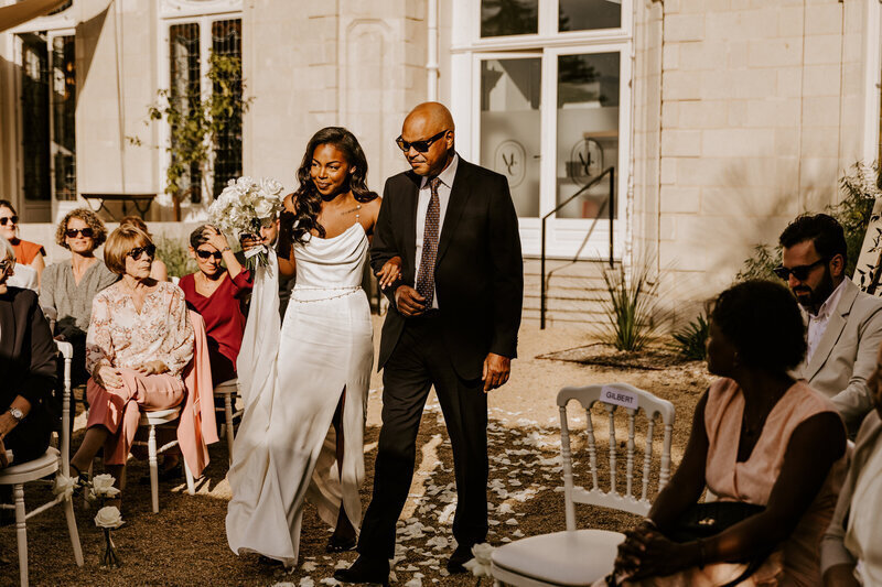 Entrée de la mariée au bras de son papa pour la cérémonie laïque sous les yeux des invités. Photo de mariage prise par Laura Termeau.