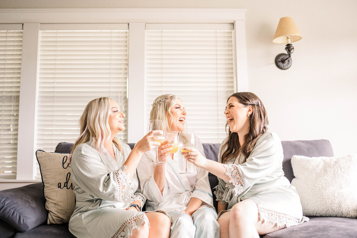 three bridesmaids cheers with mimosas while sitting on a couch on wedding morning