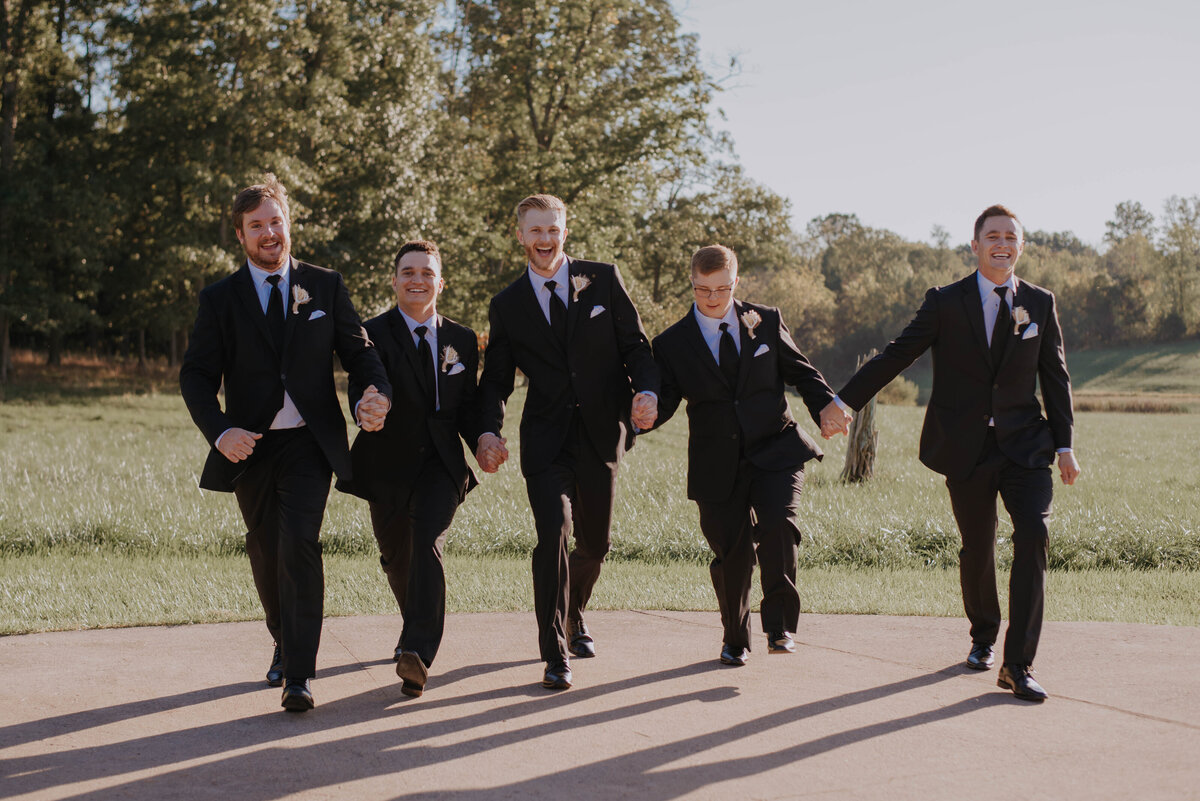 Five men in black suits and ties stroll hand in hand outdoors, exuding joy as they wander through a picturesque Leesburg, VA wedding venue. Surrounded by lush greenery and trees under a clear blue sky, their smiles shine brighter than the best wedding packages Virginia has to offer.