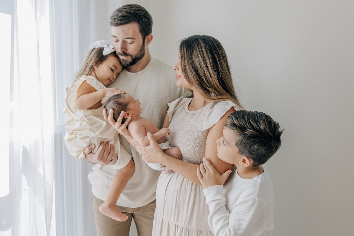 family of 5 standing by window holding newborn baby