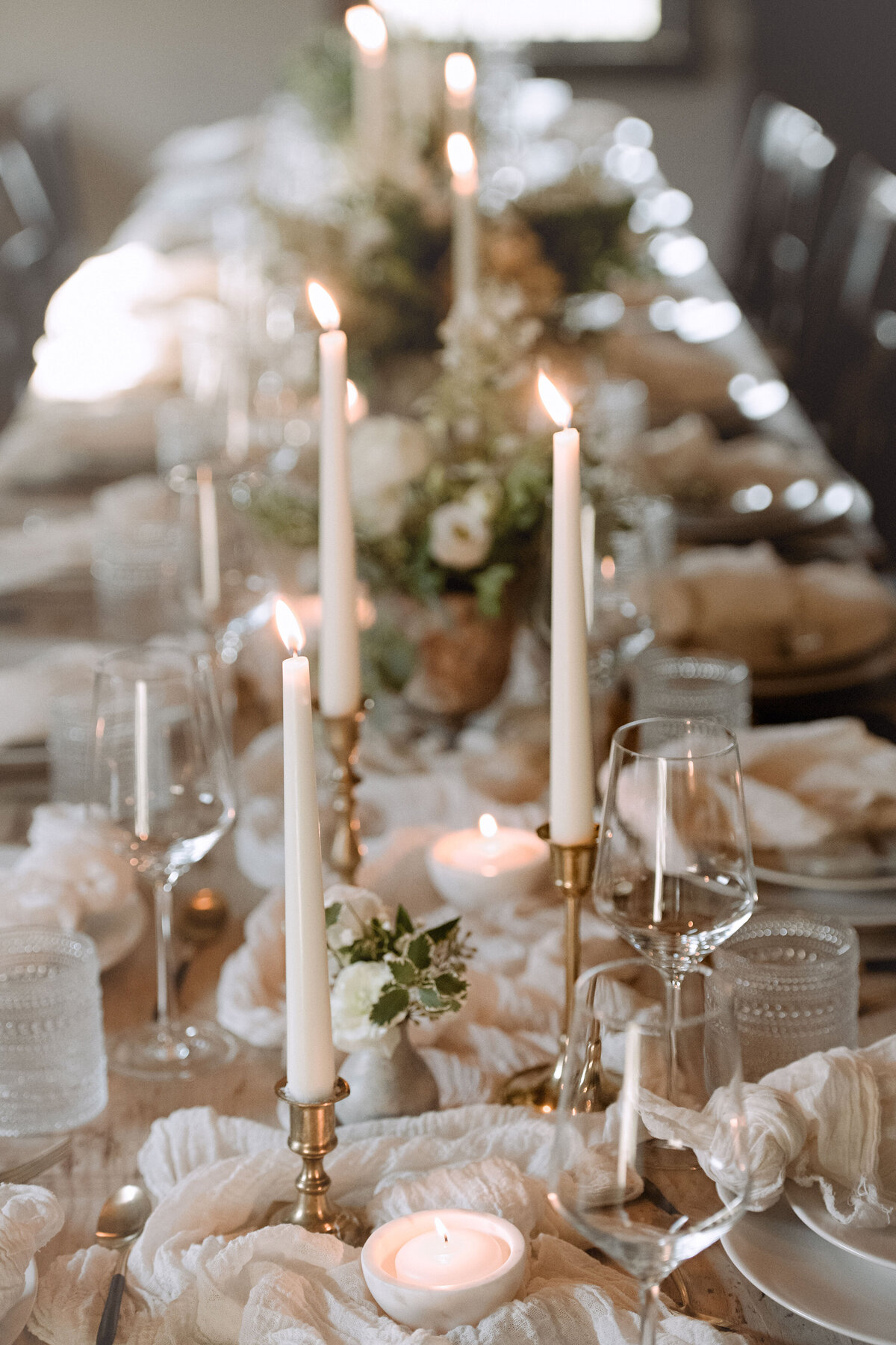 A rustic wedding reception table set with taper candles in brass holders, a gauzy table runner, and ivory linens and china