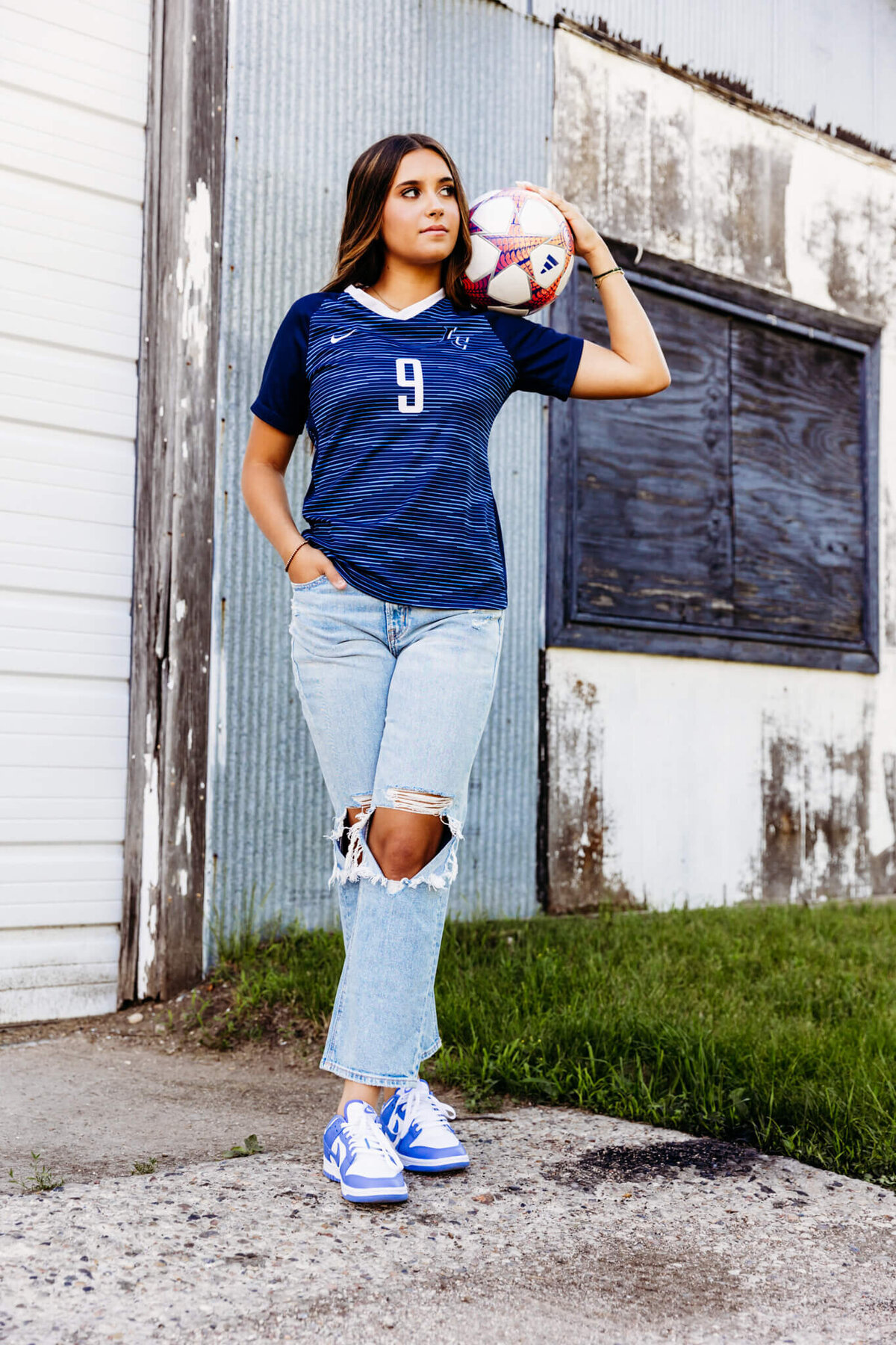 pretty teen girl in a blue striped soccer jersey standing with her legs crossed and a soccer ball on her shoulder as she poses for her senior photos