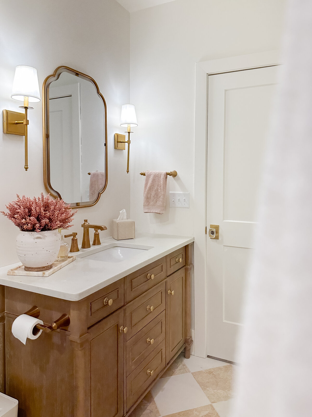 A guest bathroom features a wood vanity in a warm finish with brass hardware and sconces