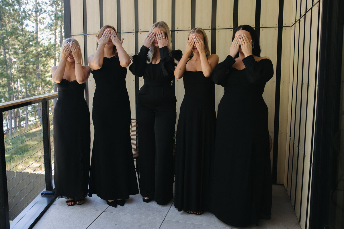 Five bridesmaid cover their eyes with their hands. They are all in different variations of black formal dresses. Behind them a is a wooden paneled wall with thick black trim. 