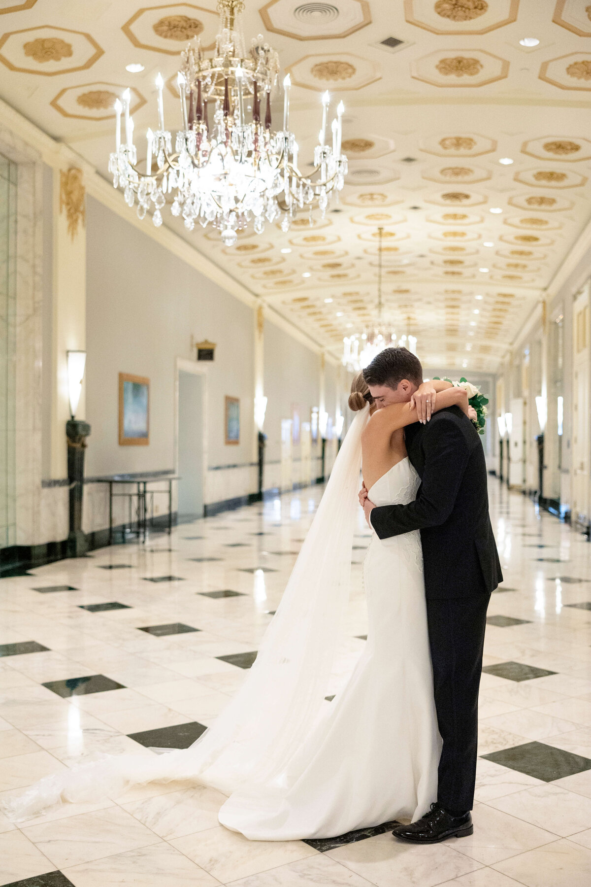 A bride and groom embrace in a grand hallway with elegant chandeliers and marble floors. The bride wears a long white dress with a veil, and the groom is in a black suit. The hallway is ornate, with framed artwork on the walls.