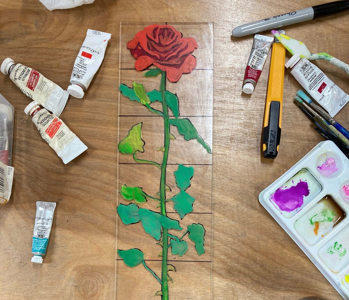 A detailed red rose monoprint painted across several plexiglass tiles, surrounded by paint tubes and tools, highlighting the setup before printing.