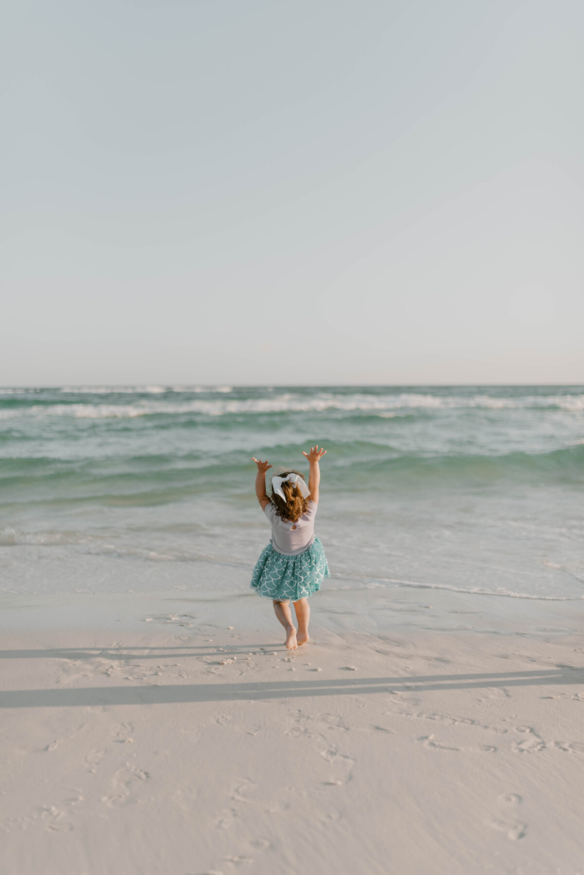 a little girl throwing her hands in the air while dancing in the florida beach sand as the waves reach her toes.