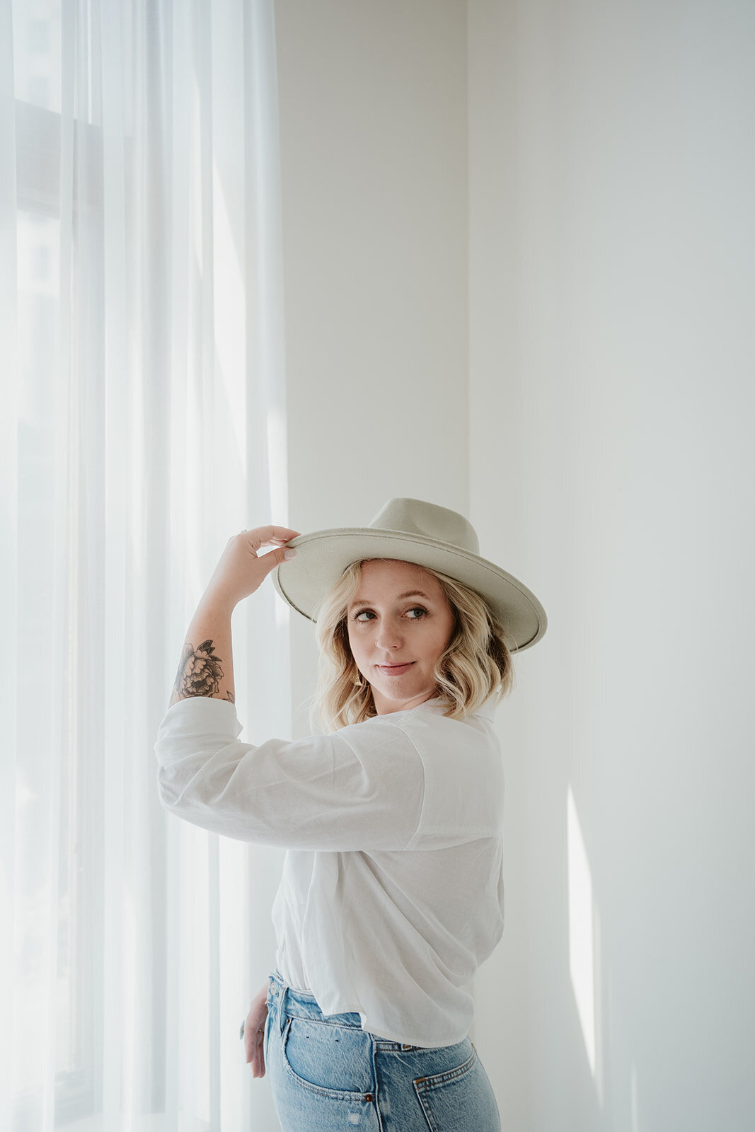 Portrait of a woman wearing a white shirt and hat posing in front of a window during a Wildher and Co branding session in downtown Kalamazoo Michigan.